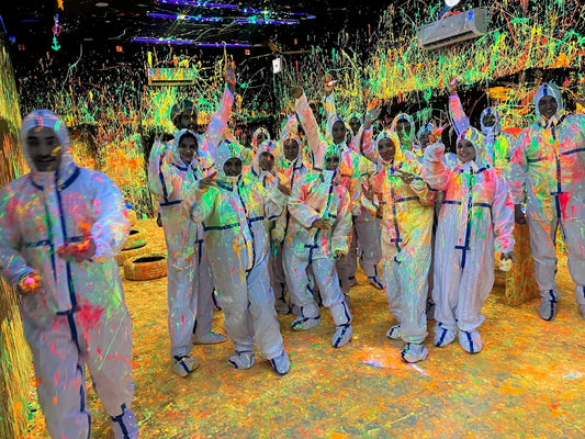 A group of young people in white protective suits are covered in neon paint splatters in a dark room. They are smiling and posing for a photo.