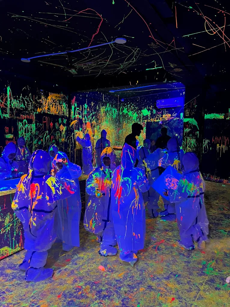 People in protective suits covered in neon paint splatters in a blacklight room.