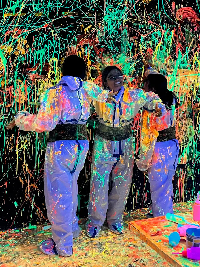 Three young people in paint-splattered suits stand in front of a black wall covered in neon paint splatters.
