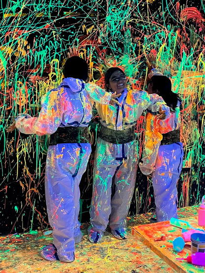 Three young people in paint-splattered suits stand in front of a black wall covered in neon paint splatters.