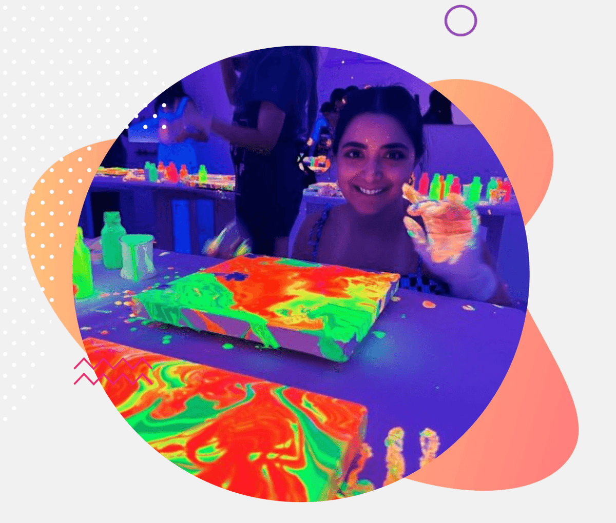 A young woman smiles while painting with neon colors under blacklights. Two canvases with vibrant, swirled paint are on a table.
