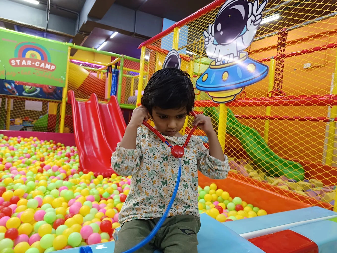 A young child plays doctor with a stethoscope in a colorful ball pit at an indoor playground.