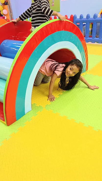 A young girl crawls through a rainbow tunnel in a colorful indoor play area.
