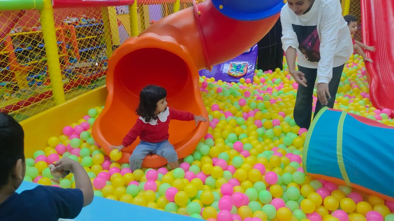 A young child sits in a ball pit with a slide, while a woman and another child are visible in the background.
