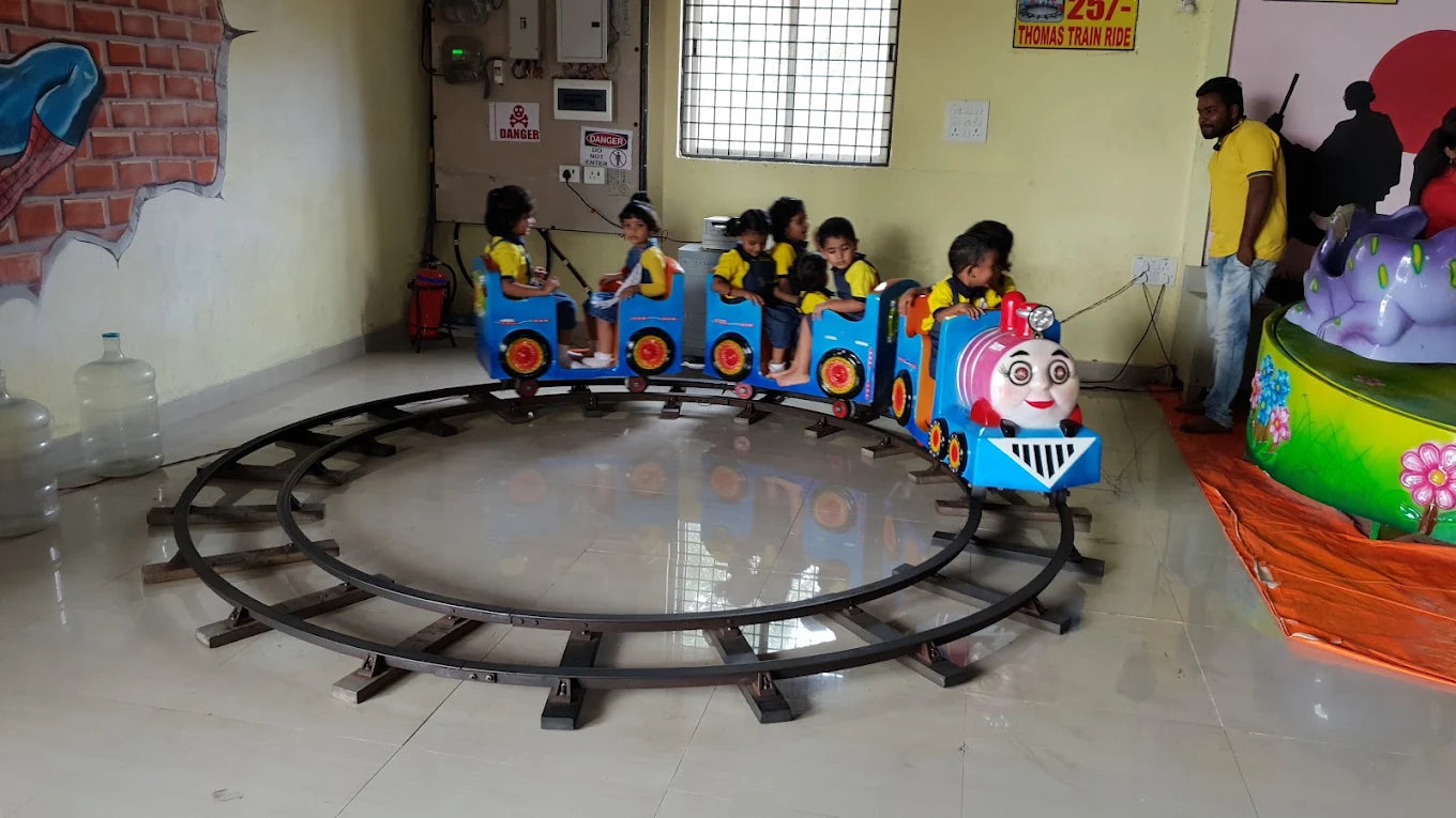 Children ride a toy train on a circular track in an indoor play area. A man supervises the ride.