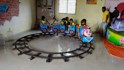 Children ride a toy train on a circular track in an indoor play area. A man supervises the ride.
