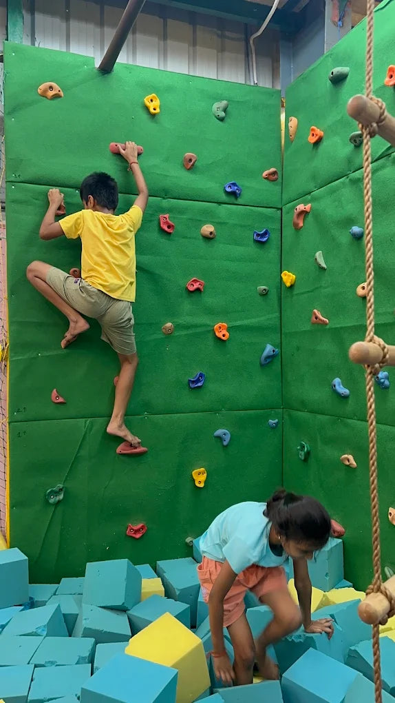Two children climb a green rock wall above a pit of blue and yellow foam blocks.