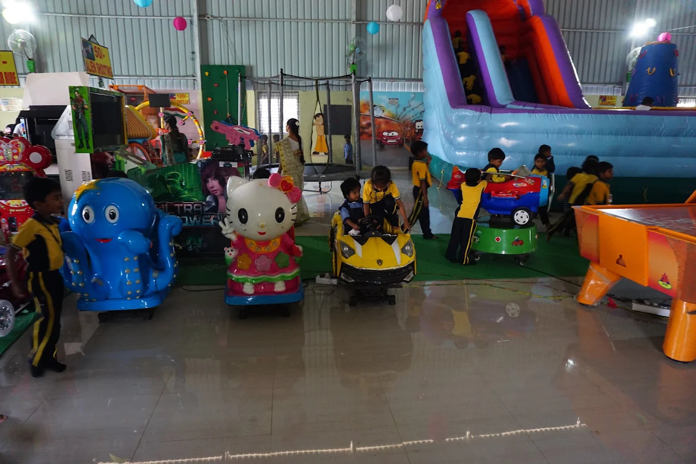 Children play on rides and games in an indoor amusement center. A large inflatable slide is visible in the background.