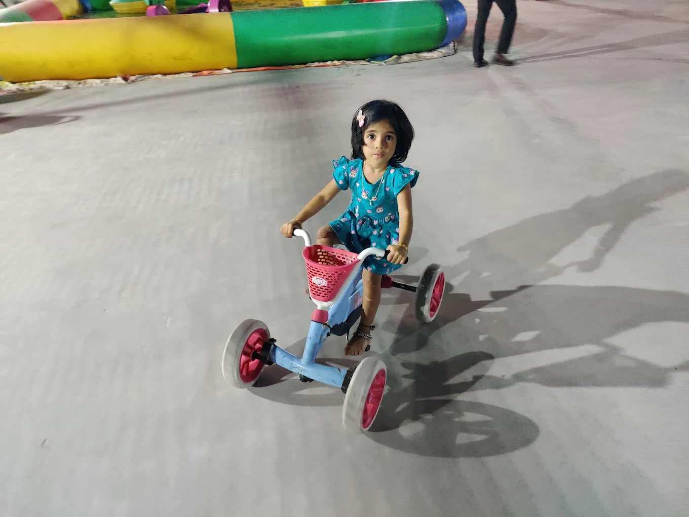 A young girl rides a blue tricycle with a pink basket on a paved surface.
