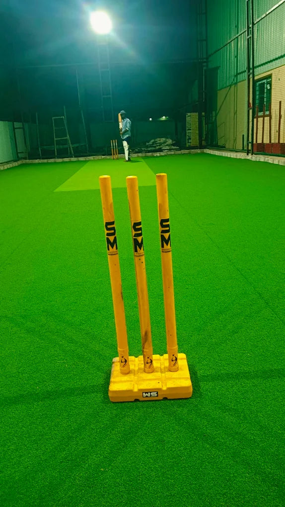 A person plays cricket on a green turf field at night, with wickets in the foreground.