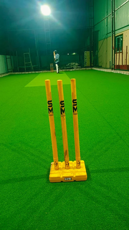 A person plays cricket on a green turf field at night, with wickets in the foreground.