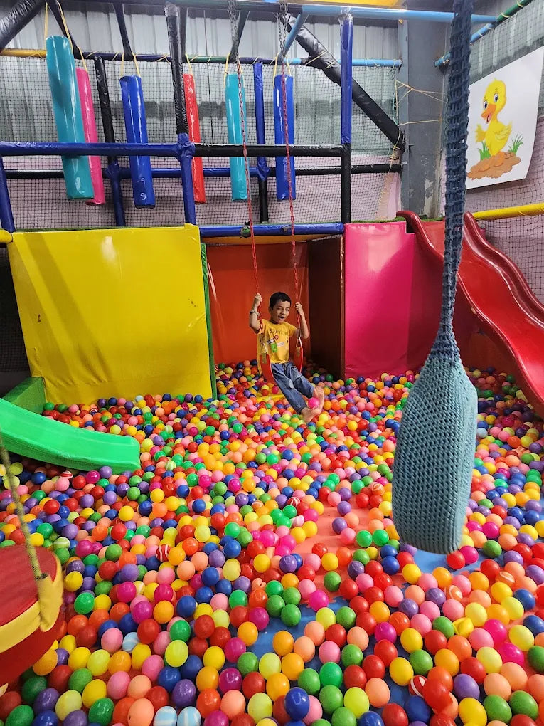 A young boy swings on a swing over a ball pit in an indoor playground.