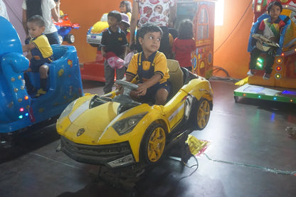 Young children ride in colorful toy cars at an indoor amusement area.