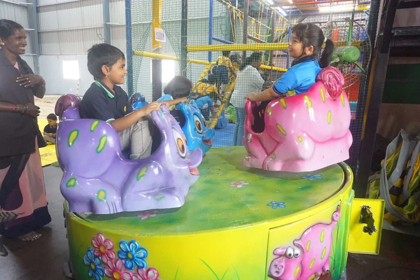 Children ride on a colorful carousel with animal-shaped seats in an indoor play area.