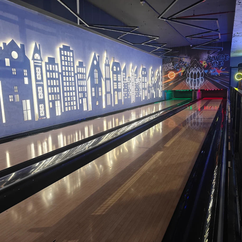 A bowling alley with illuminated city skyline art on the wall and reflections on the lanes.
