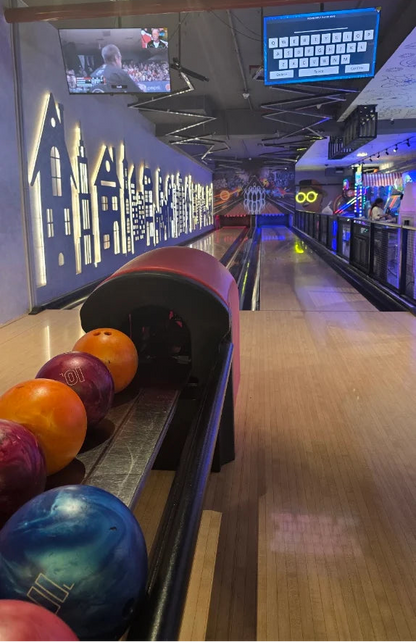 Bowling balls lined up on a lane in an arcade with people playing games in the background.