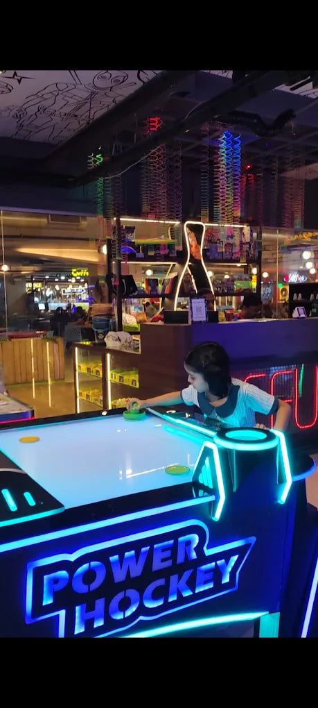 A young girl plays air hockey in a brightly lit arcade. The game table glows with blue neon lights.