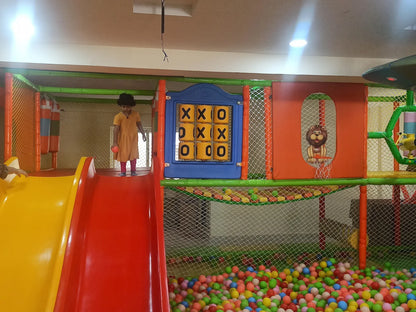 A young child stands on a slide in a colorful indoor playground with a ball pit.