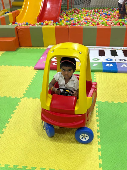 A young child sits in a toy car in a colorful indoor playground.