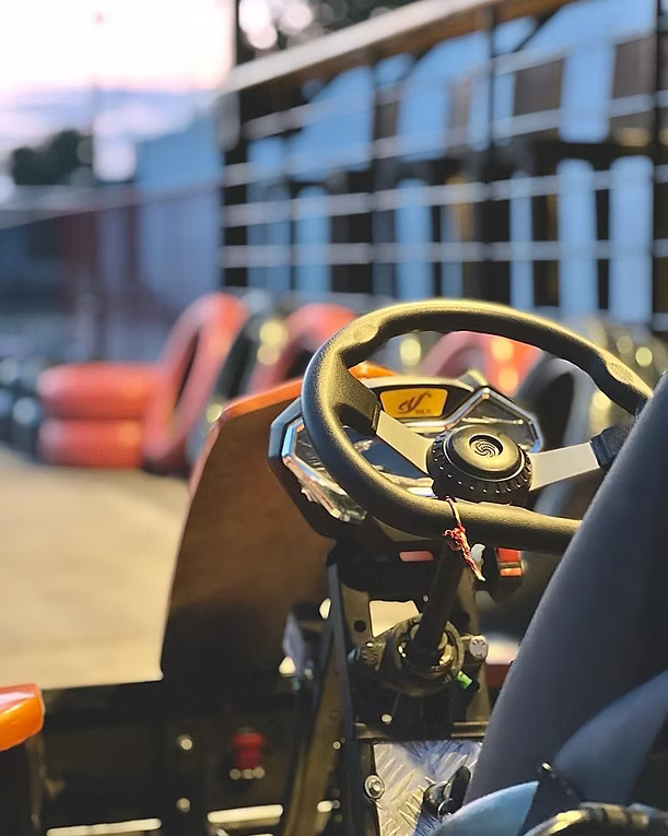 Close-up of a go-kart steering wheel and dashboard, with orange barriers and a track in the background.