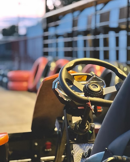 Close-up of a go-kart steering wheel and dashboard, with orange barriers and a track in the background.