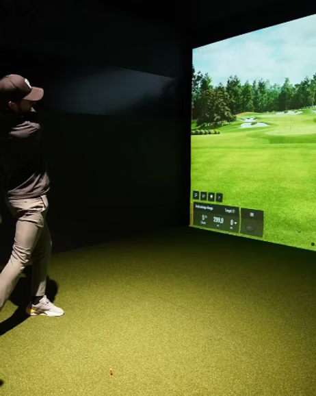 A person in athletic wear swings a golf club in a simulator room with a projected golf course.