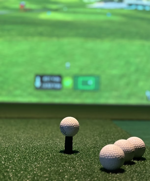 Golf balls on a tee and on the ground in front of a golf simulator screen.