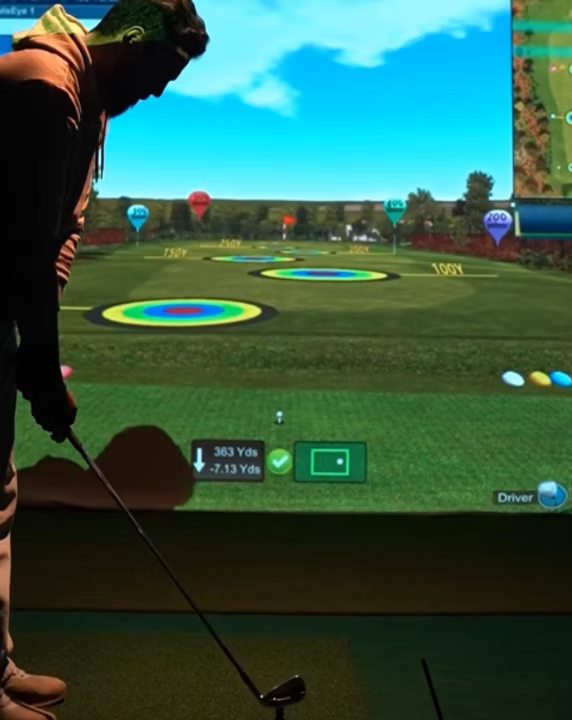 A young person practices golf on a simulator, with a virtual driving range displayed on a large screen.