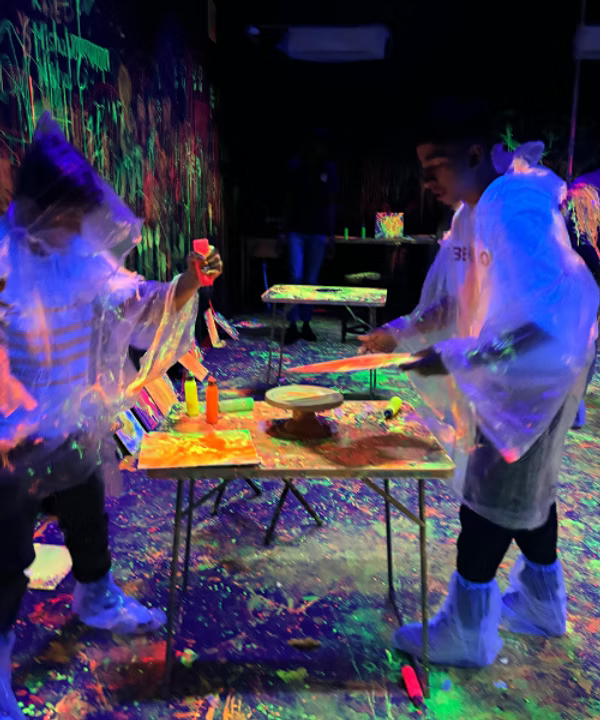 Two young people in a neon-lit room, splattered with paint, are engaged in a creative activity. They are wearing protective ponchos and shoe covers.