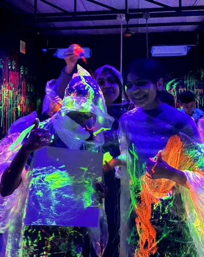 Young people in a blacklight room, covered in neon paint, hold up glowing artwork.
