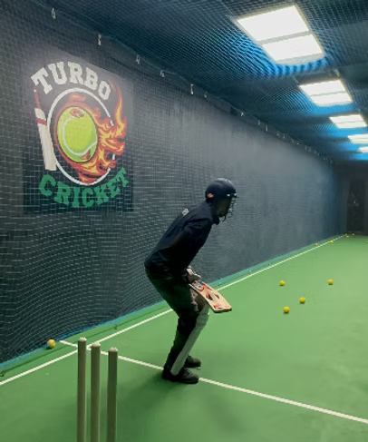 A young person in cricket gear practices in an indoor net. Several yellow balls are scattered on the green turf.