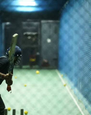 A young person bats during a cricket practice session in an indoor net. Yellow balls are scattered on the green court.