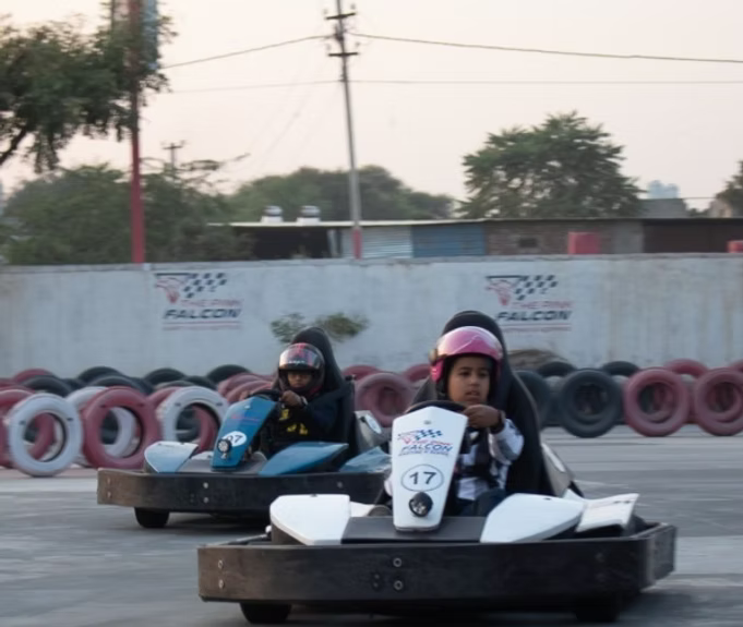 Two young children in go-karts race on a track, wearing helmets.