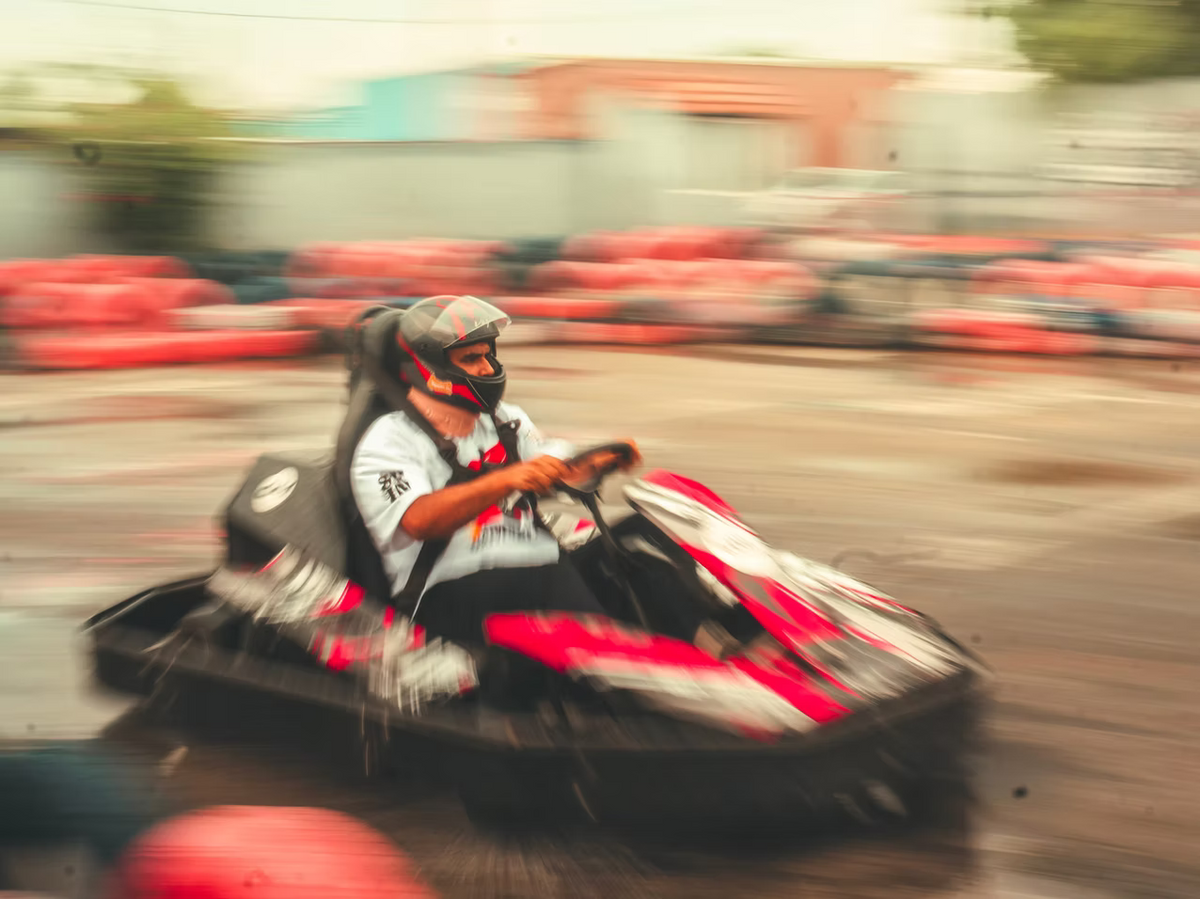 A person in a helmet races a go-kart on a track, with blurred red barriers in the background.