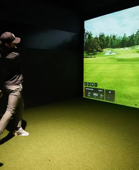 A man plays golf on a simulator, swinging at a virtual course displayed on a large screen.