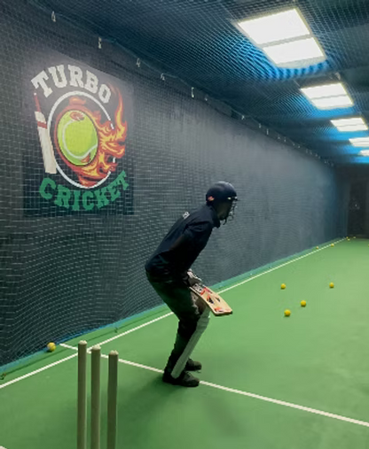 A young person in cricket gear practices in an indoor net with yellow balls scattered on the green turf.