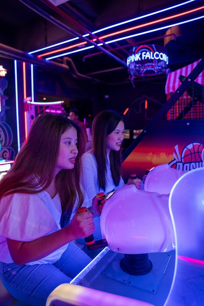 Two women playing an arcade game with neon lights and a basketball hoop in the background.