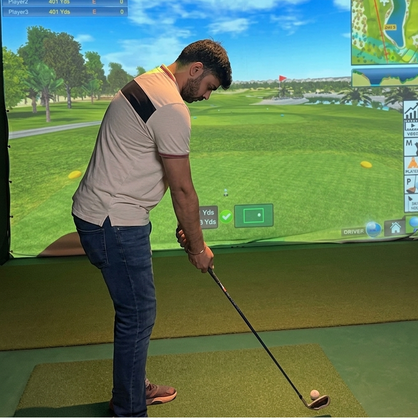 A young man plays golf on a simulator, focusing on his swing. The screen displays a virtual golf course.