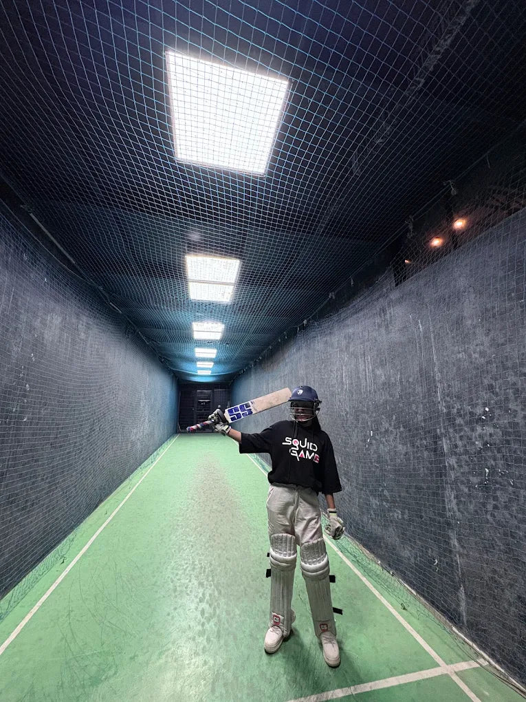 A young person in cricket gear stands in a batting cage, holding a bat.