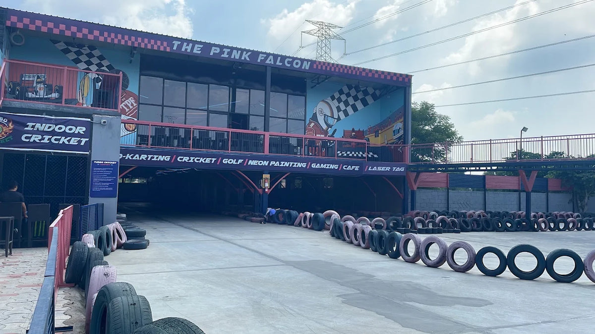 An outdoor go-kart track with a building featuring "The Pink Falcon" and various activities. Tires line the track, and a few people are visible.