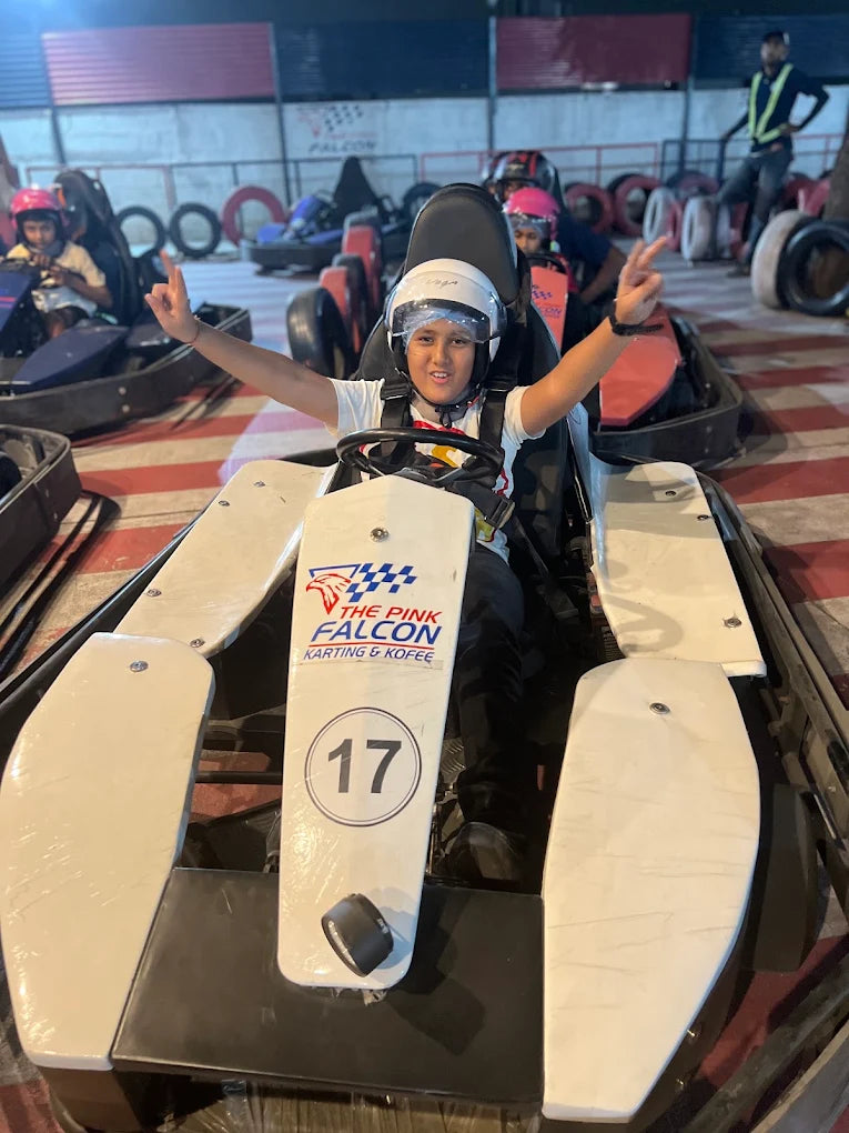 A young person in a go-kart smiles and gives a peace sign, ready to race at an indoor track.