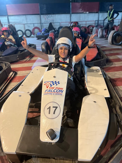 A young person in a go-kart smiles and gives a peace sign, ready to race at an indoor track.