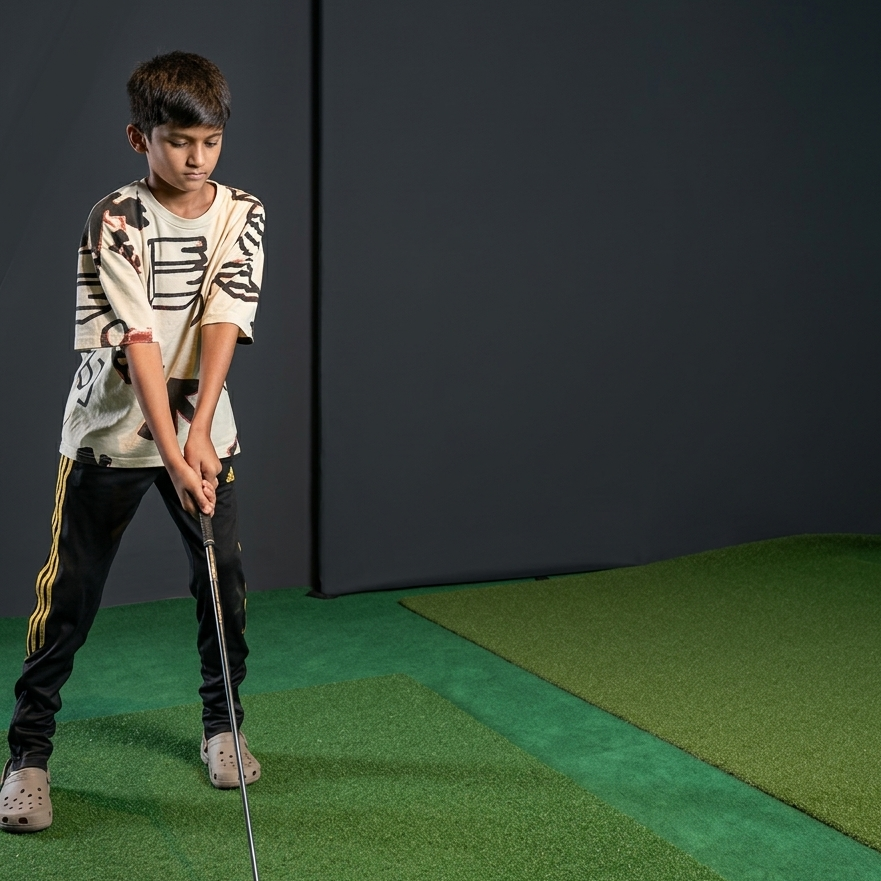 A young boy practices golf indoors on a green mat, holding a club.