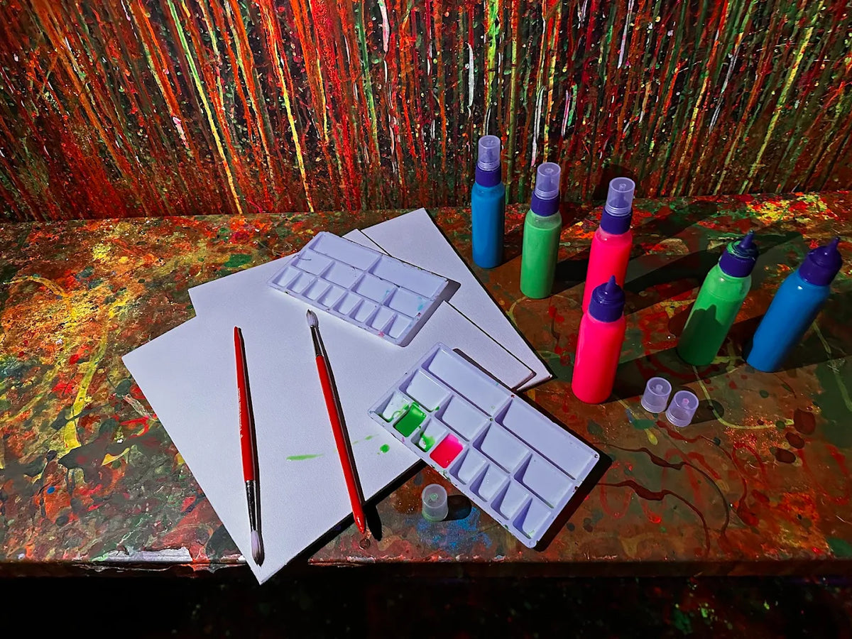 Art supplies, including paint bottles and brushes, are arranged on a colorful, paint-splattered surface.