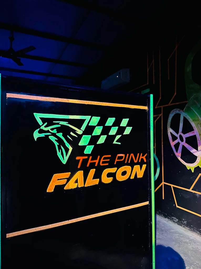 A neon sign for "The Pink Falcon" glows in a dark, possibly indoor, setting.