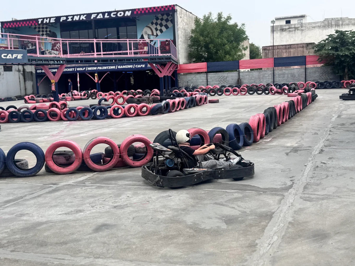 A person in a go-kart races on a track lined with colorful tires, with a building labeled "The Pink Falcon" in the background.