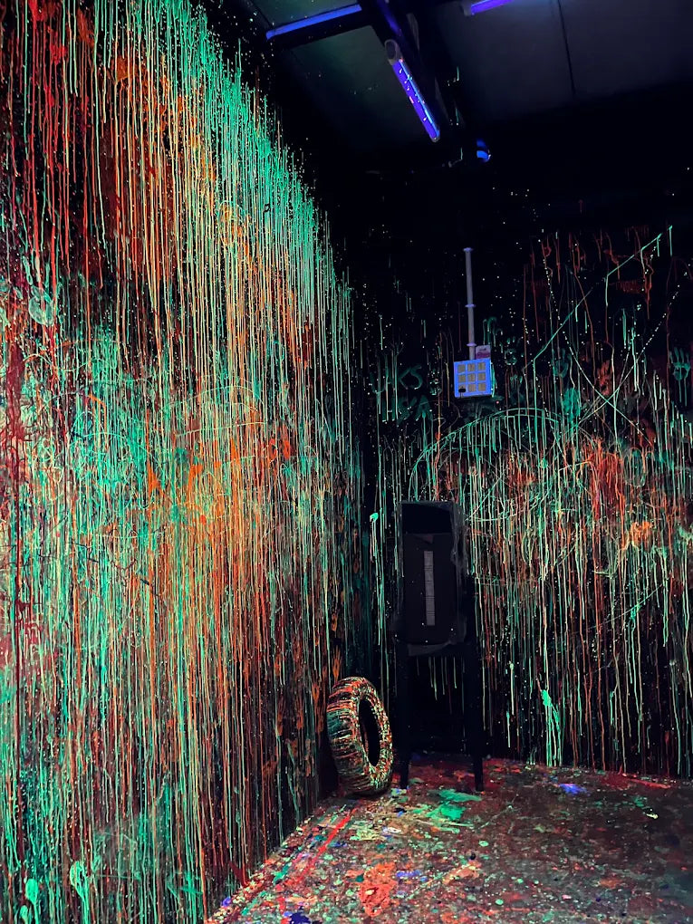 A room with walls and floor covered in neon paint drips, with a tire and a black object in the corner.