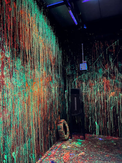 A room with walls and floor covered in neon paint drips, with a tire and a black object in the corner.