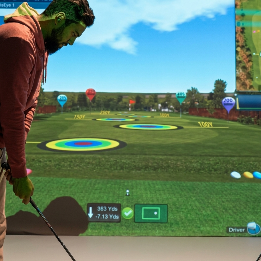 A young man plays virtual golf indoors, aiming at a projected driving range with distance markers.