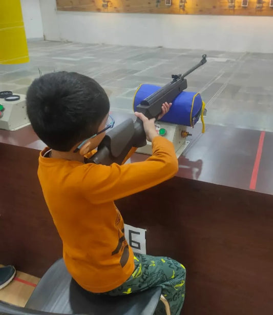 A young boy practices shooting with a rifle at a range. He is wearing an orange shirt and patterned pants.